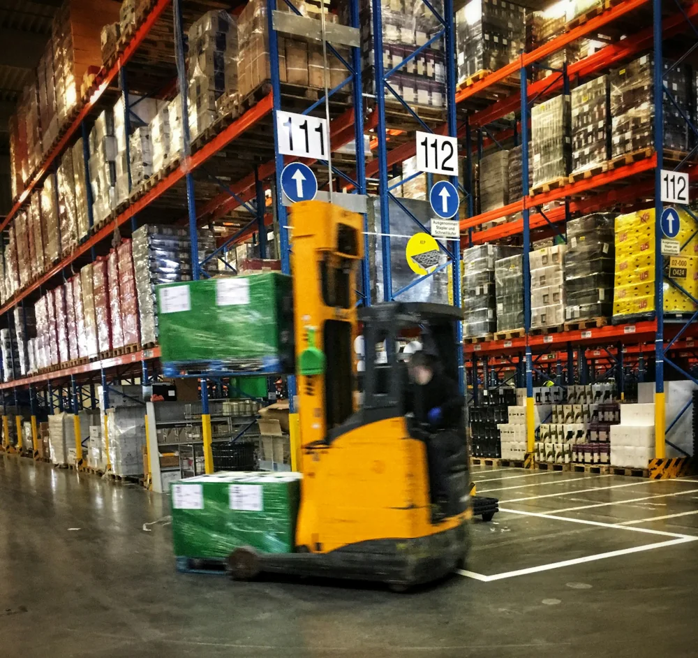 A yellow forklift moves a green-wrapped pallet past tall warehouse racks filled with packaged goods.